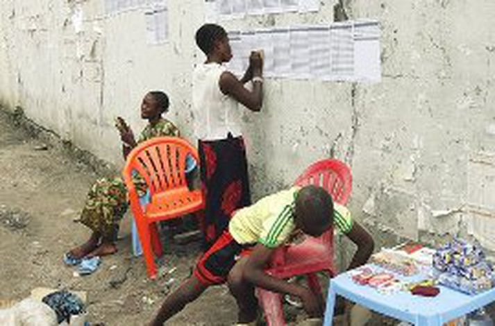 Íbúi Kinshasa leggur hér saman úrslit kjörstöðvar þar sem þau hanga utan á vegg kjörstöðvarinnar. Einnig má þar sjá framtakssaman ungling sem heldur úti sjoppurekstri á staðnum.