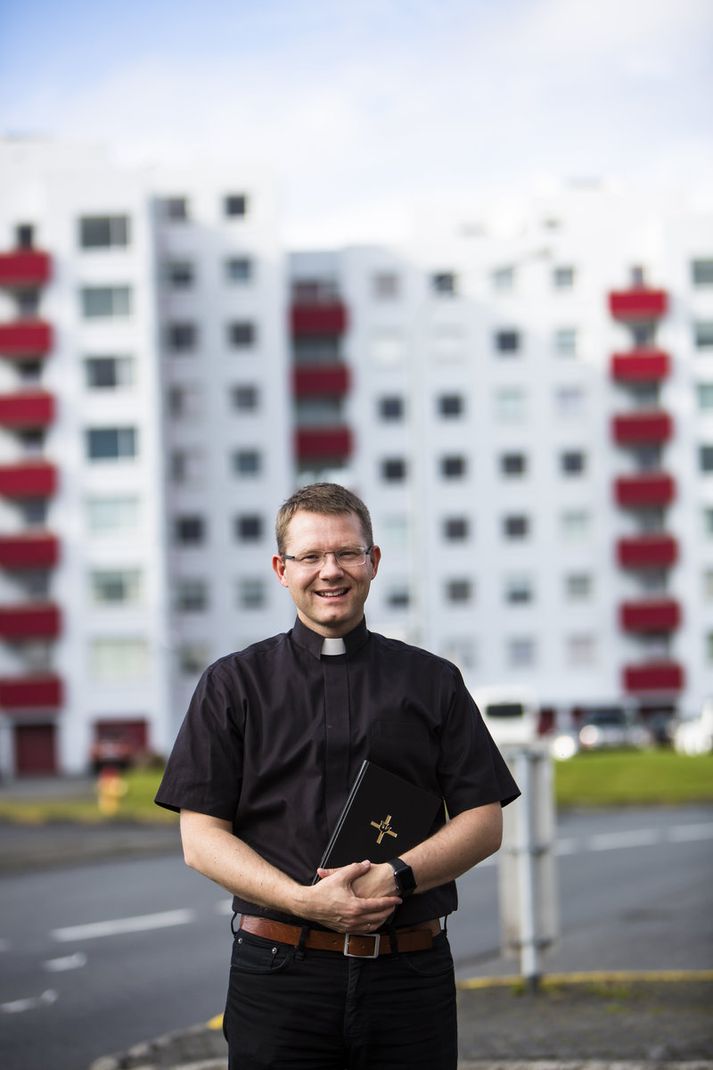 Séra Jón Ómar Gunnarsson með prestkraga og Biblíuna í hendi framan við tignarleg og glæst háhýsin við Asparfell í Efra Breiðholti.