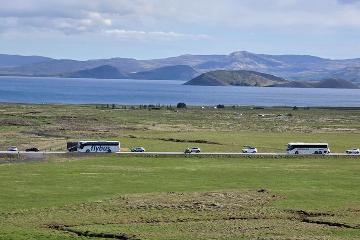 Hér má sjá tvær hópbifreiðar svífa fram úr bílaröðinni í fylgd lögreglumanns á bifhjóli. Í baksýn er má sjá Þingvallavatn. 